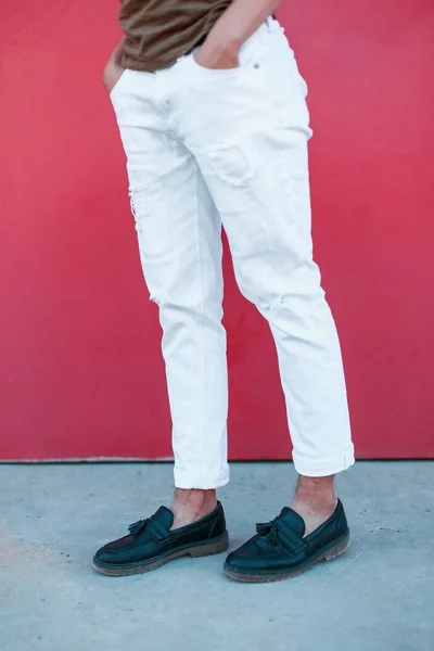 Stylový mladík stojí nedaleko kovové a zářivě růžové zdi. Kožené černé boty a stylové bílé kalhoty. Moderní sezónní Pánské boty. — Stock fotografie