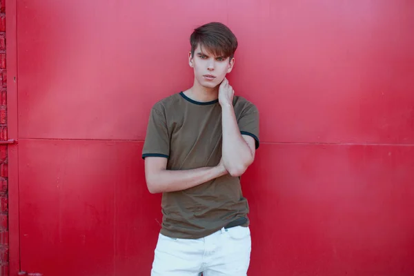 Joven guapo fresco con un peinado elegante en una camiseta verde de moda en pantalones blancos cerca de la pared de metal rosa brillante en un cálido día de verano. El tipo atractivo está descansando al aire libre. Estilo juvenil . — Foto de Stock