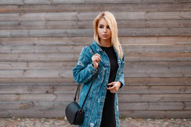 Şık bir deri çanta ile güzel genç sarışın kadın bir ahşap Vintage duvar yakın siyah bir elbise moda denim ceket. Bir yürüyüş için çekici Amerikan kız modeli. Gençlik tarzı giyim.