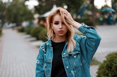Moda kentsel sevimli genç kadın bir trendy uzun Kot ceket bir parkta bir yaz gününde poz beyaz spor ayakkabı içinde bir siyah şık elbiseli. Sevimli kız açık havada dinlenme. Retro tarzı.