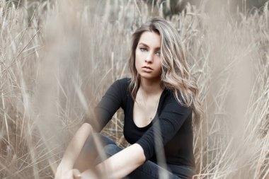 Moda siyah gömlek şık kot güzel genç kadın sarışın kuru sonbahar çim arasında bir alanda oturan istirahat. Sevimli kız modeli sıcak bir günde köyde doğa sahiptir.