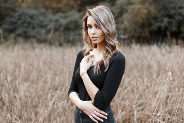 Şık kot sarı saçlı siyah şık T-shirt güzel güzel seksi genç kadın sıcak bir sonbahar gününde bir alanda pozlar. Modern sevimli kız modeli açık havada yürür.