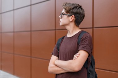 Bir sırt çantası ile kahverengi bir tişört şık güneş gözlüğü yakışıklı genç moda hipster adam bir yaz gününde modern bir binanın yakınında pozlar. Çekici modern Trendy genç.