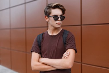 Bir spor sırt çantası ile bir yaz t-shirt içinde güneş gözlüğü çekici genç hipster adam bir Vintage bina yakın güneşli bir günde bir sokakta duruyor. Yakışıklı moda model adam şehirde dinleniyor.