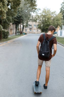 Atletik genç yakışıklı hipster adam trendy yaz giysileri siyah bir sırt çantası ile snowboard sürmek. Şık adam yaz günü sahiptir. Büyük hobi. Arkadan görüntülemek.