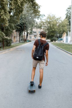 Bir hipster t-shirt bir sırt çantası ile yaz şort modern genç yakışıklı adam parkta bir kaykay yol boyunca sürme. Amerikalı adam şehir etrafında seyahat ediyor. Arkadan görünüm.