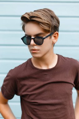 Genç şık yakışıklı hipster adam şık kahverengi tişört siyah güneş gözlüğü, son moda saç modeli ile bir ahşap mavi duvar poz. Amerikan modern adam dinleniyor. Yaz günü.