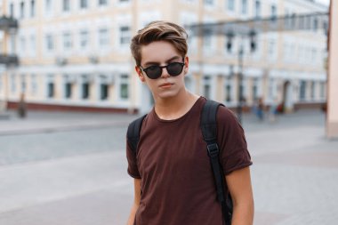Şık bir saç modeli ile kahverengi bir trendy t-shirt içinde güneş gözlüğü yakışıklı genç adam hipster Vintage binaların arka planda sıcak bir yaz gününde şehir boyunca yürür. Modern adam seyahat ediyor.