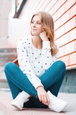 Spor ayakkabıları şık kot beyaz bir kazak neşeli güzel Avrupa Genç kadın oturur ve sokakta modern tuğla binanın yakınında rüyalar. Sevimli sarışın kız bir tatil sahiptir.