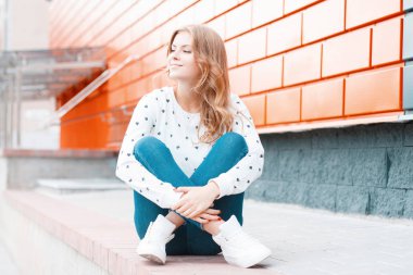 Şık kıyafetler güzel genç mutlu kadın bir yaz gününde tuğla modern duvar yakın şehirde asfalt üzerinde oturuyor. Güzel moda model kız büyük bahar hava sahiptir.