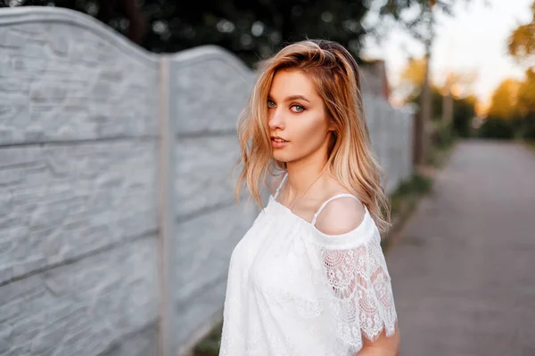 Hermosa mujer rubia joven europea en una blusa elegante de encaje blanco está descansando al aire libre al atardecer. Feliz modelo chica atractiva disfrutar de vacaciones de verano. Ropa de mujer de estilo primavera . —  Fotos de Stock