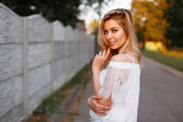 Jovem atraente positivo em uma blusa de renda elegante posando ao ar livre perto da cerca em um dia ensolarado de verão. Modelo menina feliz muito elegante em uma caminhada . — Fotografia de Stock