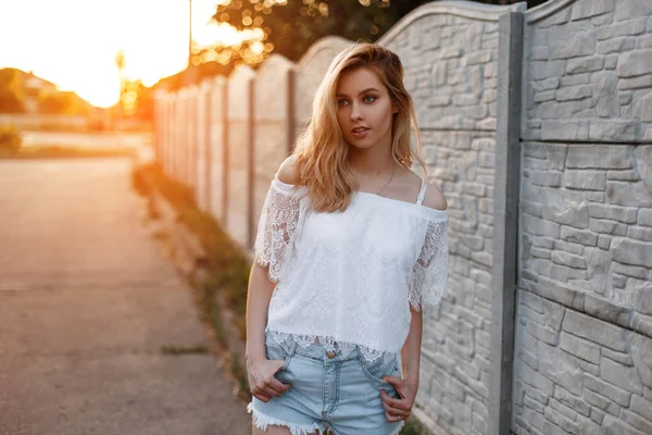 Bastante elegante jovem loira em um top de renda branca vintage em shorts jeans elegantes posando perto da cerca ao pôr do sol. Menina feliz europeia gosta de uma caminhada. Estilo de verão . — Fotografia de Stock