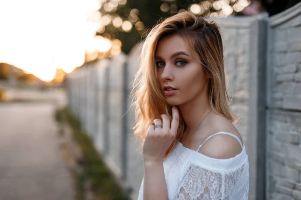 Porträt einer hübschen europäischen jungen blonden Frau mit blauen Augen und natürlichem Make-up in einer stylischen weißen Spitzenbluse in der Nähe eines Vintage-Zauns bei Sonnenuntergang. Nettes Mädchen auf einem Spaziergang im Sommer. — Stockfoto