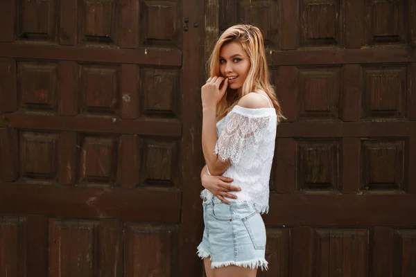 Hübsche, fröhliche junge blonde Frau mit einem süßen Lächeln in einer trendigen weißen Spitzenbluse in sommerlichen Jeanshosen posiert neben einer alten Holztür. europäisch modern girl model genießen großartiges wochenende. — Stockfoto