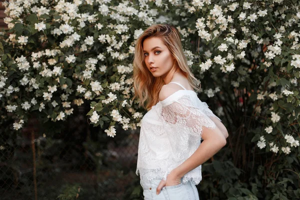 Mulher loira bonita muito jovem em uma camiseta de renda branca na moda em shorts de ganga elegantes perto de um arbusto florido com flores brancas. Modelo europeu menina desfrutando de um dia quente de primavera na natureza . — Fotografia de Stock