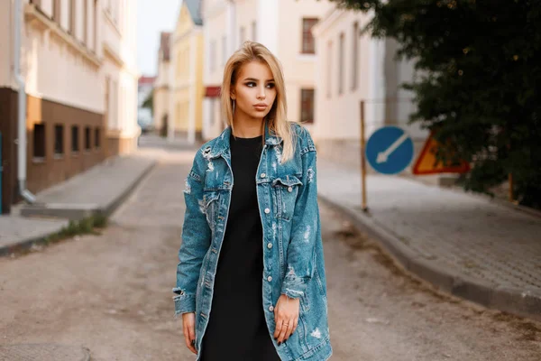 Eleganta urbana ganska ung kvinna i en svart snygg klänning i en fashionabla lång jeansjacka poserar i en stad på gatan i närheten vintage byggnader. Söt flicka utomhus. Våren stil damkläder. — Stockfoto