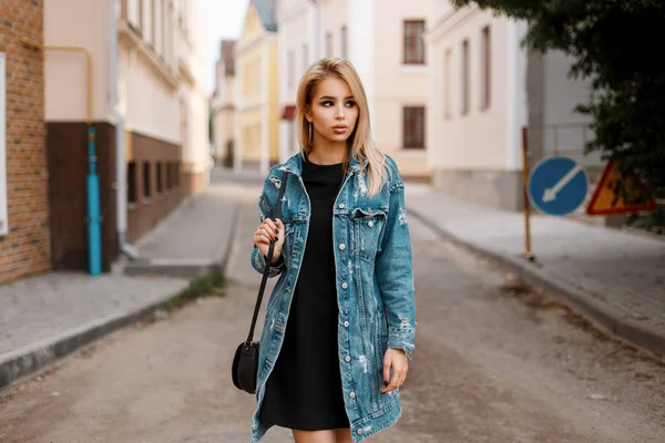 Carino bella giovane donna in un abito nero elegante con una borsa alla moda in una giacca di denim lungo alla moda in posa in una città sulla strada vicino a edifici vintage. Americano ragazza modello all'aperto . — Foto Stock