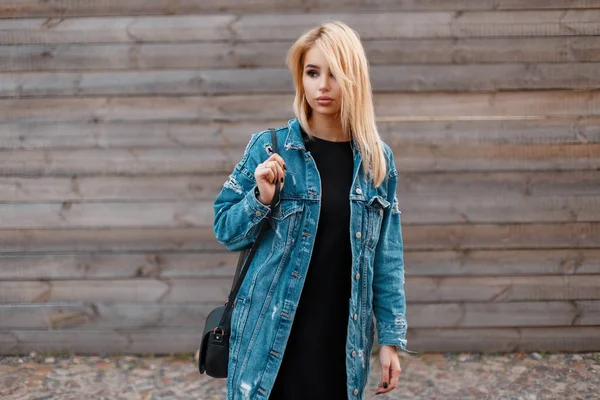 Mulher loira jovem na moda moderna com bolsa preta na moda em uma jaqueta jeans longa e elegante em um vestido preto está de pé perto da parede vintage de madeira ao ar livre. Menina muito glamour gosta do fim de semana . — Fotografia de Stock