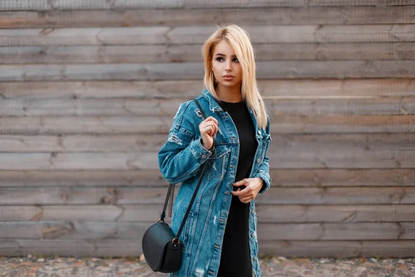 Schöne junge blonde Frau mit einer stilvollen Lederhandtasche in einer modischen Jeansjacke in einem schwarzen Kleid in der Nähe einer hölzernen Vintage-Wand. attraktive amerikanische Mädchen Modell für einen Spaziergang. Kleidung im Jugendstil. — Stockfoto