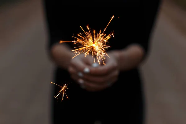 Woman in a black T-shirt celebrates the holiday with amazing sparklers. Female hands with sparkler close-up. Focus on bright fire. — Stock Photo, Image