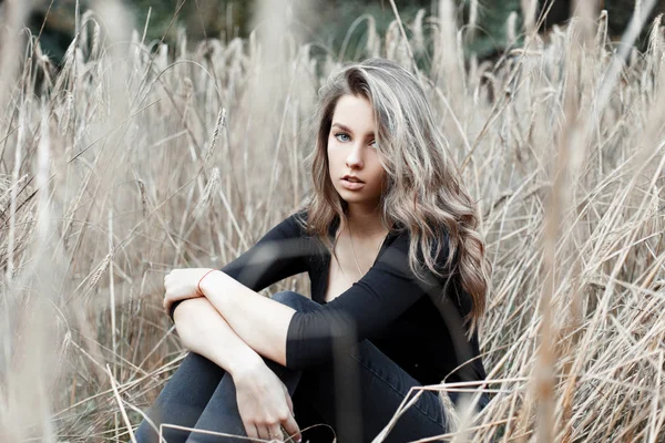 Linda jovem mulher bonita loira em jeans elegantes em uma camisa preta na moda está descansando sentado em um campo entre a grama seca do outono. Bonito modelo menina goza da natureza na aldeia em um dia quente . — Fotografia de Stock