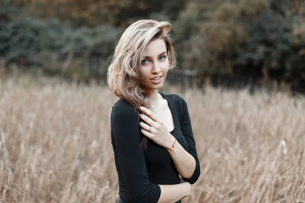 Muito atraente jovem mulher com olhos azuis com um sorriso bonito em uma camiseta preta elegante vintage está descansando em um campo perto das árvores ao ar livre na aldeia. Menina rural em um passeio no dia de outono . — Fotografia de Stock