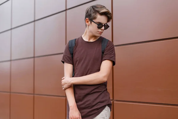 Amerikai csípő fiatal férfi egy divatos frizura napszemüveg divatos ruhákat egy fekete hátizsák jelent közel egy falat. Jóképű tinédzser fiú utazik a város körül egy nyári napon — Stock Fotó