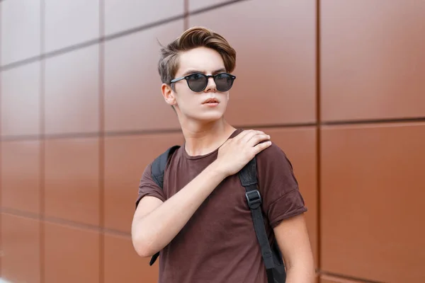 Jeune homme hipster élégant américain dans un t-shirt marron élégant en lunettes de soleil sombres avec une coiffure à la mode avec un sac à dos noir à la mode se tient près du mur. Un beau mec moderne. Style d'été . — Photo