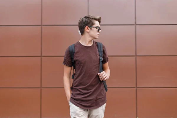 Jeune homme hipster beau dans des lunettes de soleil sombres et élégantes dans des vêtements d'été à la mode avec un sac à dos pose près du mur brun dans la ville un jour d'été. Américain touriste gars voyage autour de la ville . — Photo