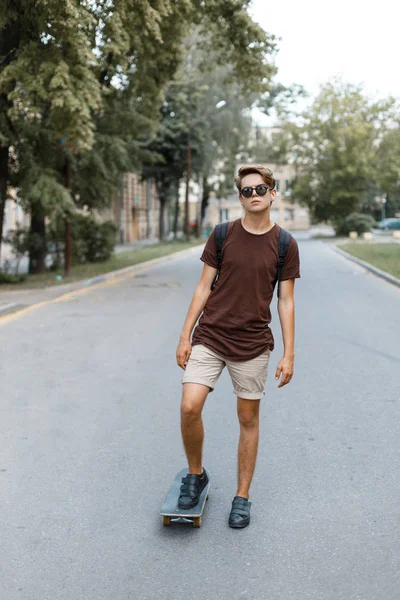 Atraktivní cool mladých bokovky muž sluneční brýle v stylové tričko v létě šortky v teniskách, ježdění na skateboardu v parku na teplý letní den. Fešák cestuje po celém městě. — Stock fotografie