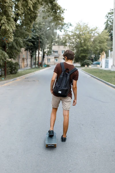 Jeune homme moderne en short d'été avec un sac à dos dans un t-shirt hipster roule le long de la route sur une planche à roulettes dans le parc. Un Américain fait le tour de la ville. Vue de l'arrière . — Photo