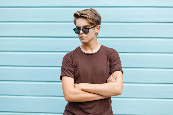 Un tânăr hipster elegant, cu o coafură la modă în ochelari de soare întunecați, într-un tricou maro la modă, pozând lângă un perete albastru din lemn într-o zi caldă de vară însorită. Bărbat american . — Fotografie, imagine de stoc