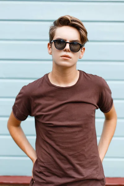 La modă tânăr hipster om modern în ochelari de soare întunecați în tricou de vară la modă, cu coafură elegantă stă lângă peretele de lemn albastru de epocă. Frumos tip în aer liber. Stilul de vară . — Fotografie, imagine de stoc