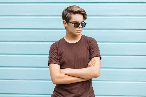 American tânăr hipster modern în ochelari de soare eleganți în tricou maro la modă stă lângă o casă albastră din lemn de epocă din oraș într-o zi de vară. Frumos tip în vacanţă . — Fotografie, imagine de stoc