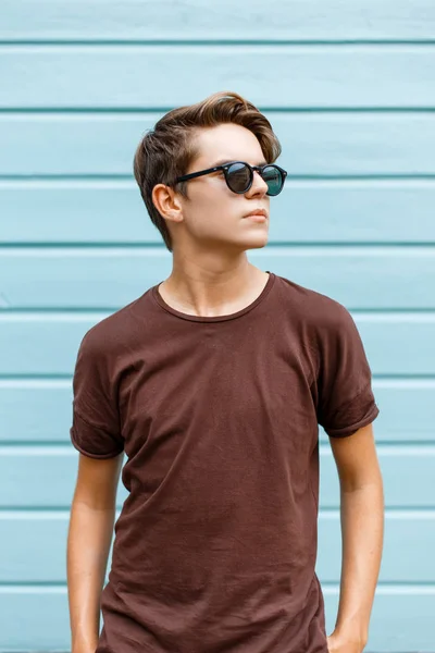 Elegante joven hombre hipster en gafas de sol con estilo en una camiseta de verano está de pie al aire libre en un día de verano cerca de una casa de madera vintage. Guapo adolescente chico está descansando en la ciudad . — Foto de Stock