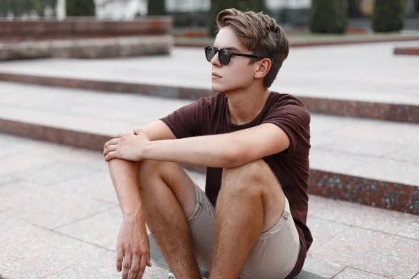 Homem jovem hipster em óculos de sol pretos com um penteado elegante em uma camiseta marrom em shorts de verão senta-se nos degraus vintage na rua. Moderno bonito cara elegante ao ar livre. Estilo de verão . — Fotografia de Stock