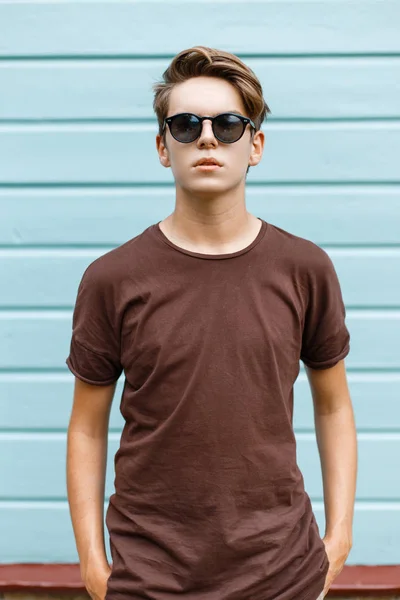 Joven hipster americano con estilo en una camiseta marrón de moda en gafas de sol negras con un peinado de moda en la calle cerca de un edificio de madera vintage. El tipo moderno a la moda al aire libre el día de verano . — Foto de Stock