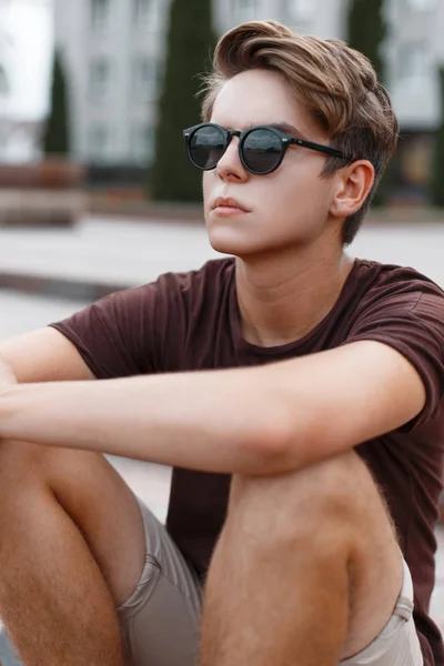 Atractivo joven hipster hombre en pantalones cortos ligeros en gafas de sol con estilo con un peinado elegante en una camiseta de moda se sienta en los escalones vintage de la ciudad. Un americano urbano al aire libre. Moda de verano . — Foto de Stock