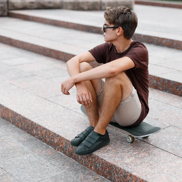 Américain jeune homme cool hipster avec coiffure dans des lunettes de soleil élégantes en été t-shirt en short en baskets assis sur un skateboard.Beau gars moderne se repose sur les marches vintage un jour d'été — Photo