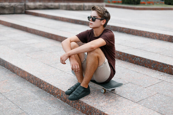 Young handsome hipster man in stylish sunglasses in stylish t-shirt in trendy shorts in sneakers resting on a skateboard in the city on a summer day. American modern guy on vacation.