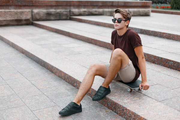 Attractive young hipster man in dark sunglasses in summer fashionable clothes in sneakers sits on a skateboard in the city on a warm summer day. Sports guy teenager.
