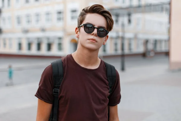 Sérieux beau jeune homme hipster dans un t-shirt à la mode avec une coiffure élégante avec un sac à dos est debout dans la rue par une chaude journée d'été. gars attrayant profiter d'une promenade dans la ville . — Photo