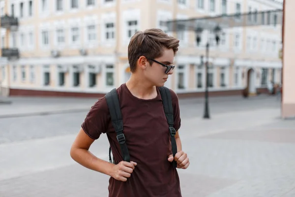 Jeune homme hipster moderne et beau dans des lunettes de soleil sombres et élégantes dans des vêtements d'été à la mode avec un sac à dos se promène dans la ville un jour d'été. Américain touriste gars voyage autour de la ville . — Photo