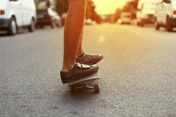 Man rider en skateboard genom staden vid solnedgången. Benen på en skateboard närbild. Mäns ben i trendiga sneakers på en skateboard på en sommar dag. — Stockfoto