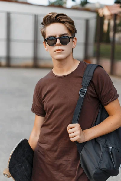 Joven hombre hipster americano en gafas de sol de moda negro en una camiseta de moda con una mochila deportiva negra con un monopatín de pie en la cancha de baloncesto. Guapo chico es relajante al aire libre . —  Fotos de Stock