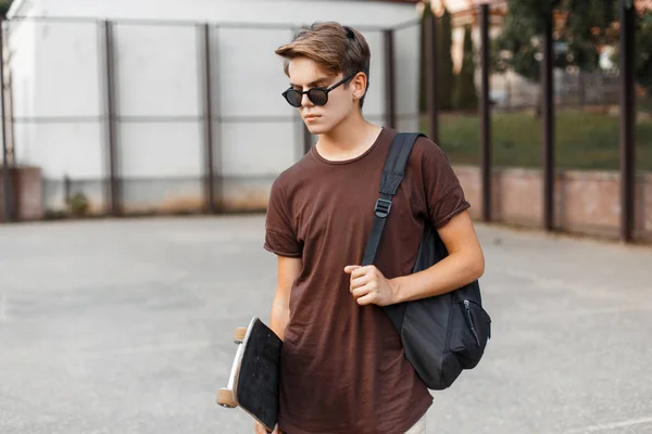 Homem hipster jovem americano em óculos de sol elegantes em uma camiseta de verão com uma mochila preta desportiva com um skate fica em um estádio em um dia quente de verão. cara legal adolescente descansando ao ar livre . — Fotografia de Stock