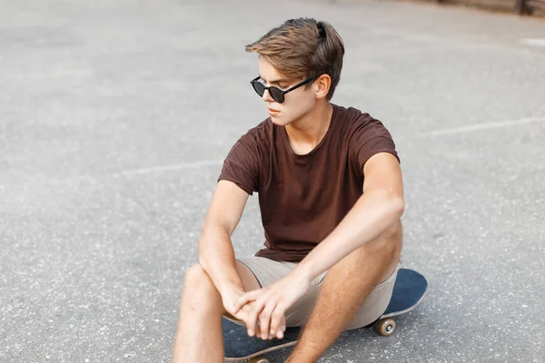 Jeune beau mâle hipster dans des lunettes de soleil à la mode en t-shirt élégant repose sur une planche à roulettes sur le terrain de basket-ball un jour d'été. Américain adolescent gars aime le week-end . — Photo