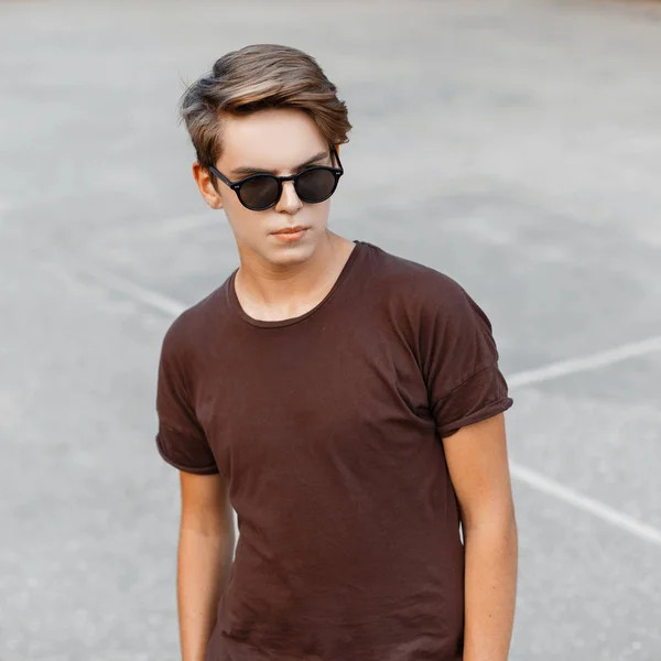 Guapo y elegante joven hipster hombre con un peinado elegante en una camiseta marrón de moda en gafas de sol de moda en la cancha de baloncesto. chico americano adolescente para un paseo verano día . — Foto de Stock