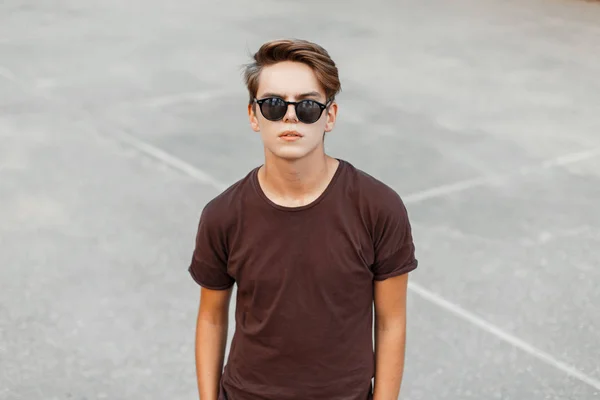 Urban hombre hipster joven de moda en elegante camiseta marrón con peinado en gafas de sol negras de moda se encuentra en el estadio. Hombre guapo americano al aire libre verano. Estilo de calle . — Foto de Stock